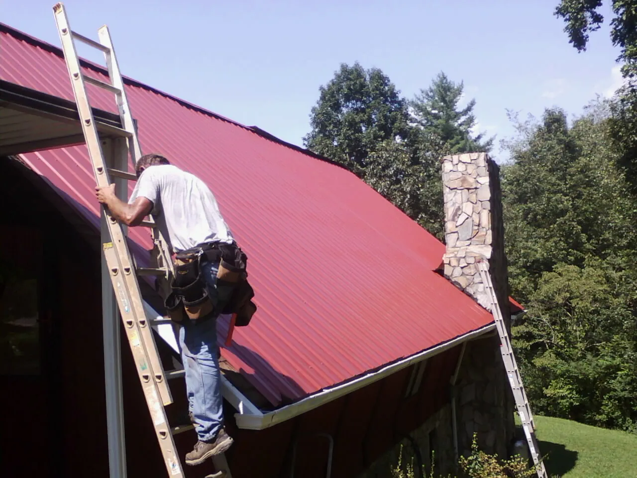 Roofer on a ladder beside a red metal roof and stone chimney — Soffit & Fascia Repair by Roofing Brothers in Claremore